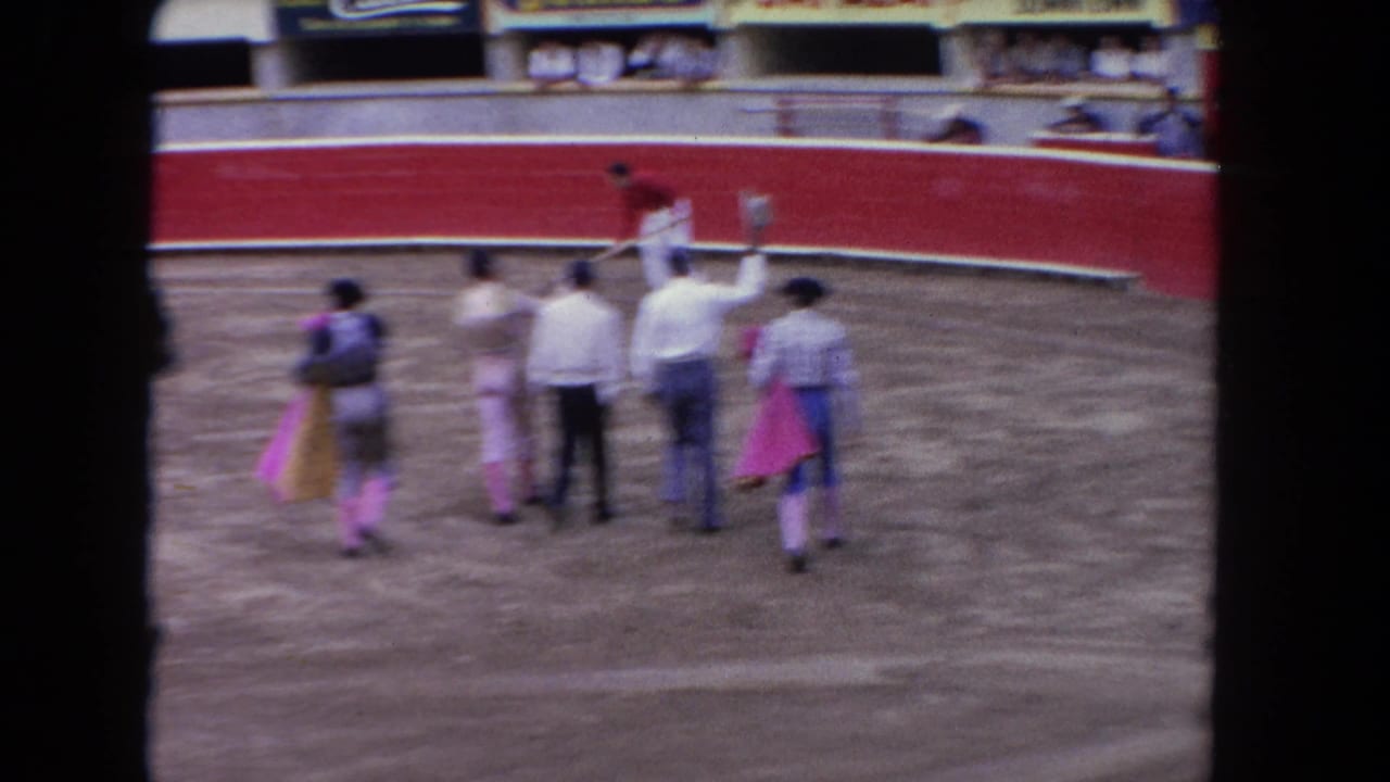 Bullfighters and officials mid-ring in a corrida arena circa 1962; red barrera wall and partial grandstand advertising signage visible behind.