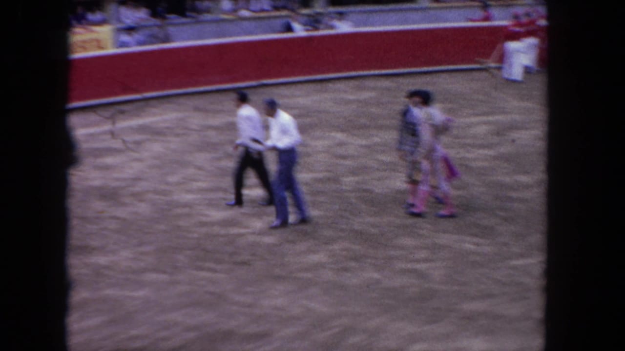 An official in a white shirt and a matador in an ornate costume cross the dirt arena; the red barrier wall and light-colored banners are visible in the background, circa 1962.