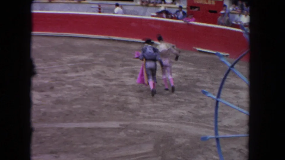 Two matadors, one carrying a pink cape, stand together in the arena with spectators visible behind the red barrier and a blue arc structure in the foreground, circa 1962 Los Angeles.