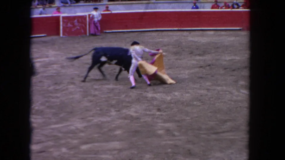 Close bullfighting action, circa 1962: matador in gold-toned traje de luces executes a pass as a dark bull moves through; red barrera and spectators visible behind.