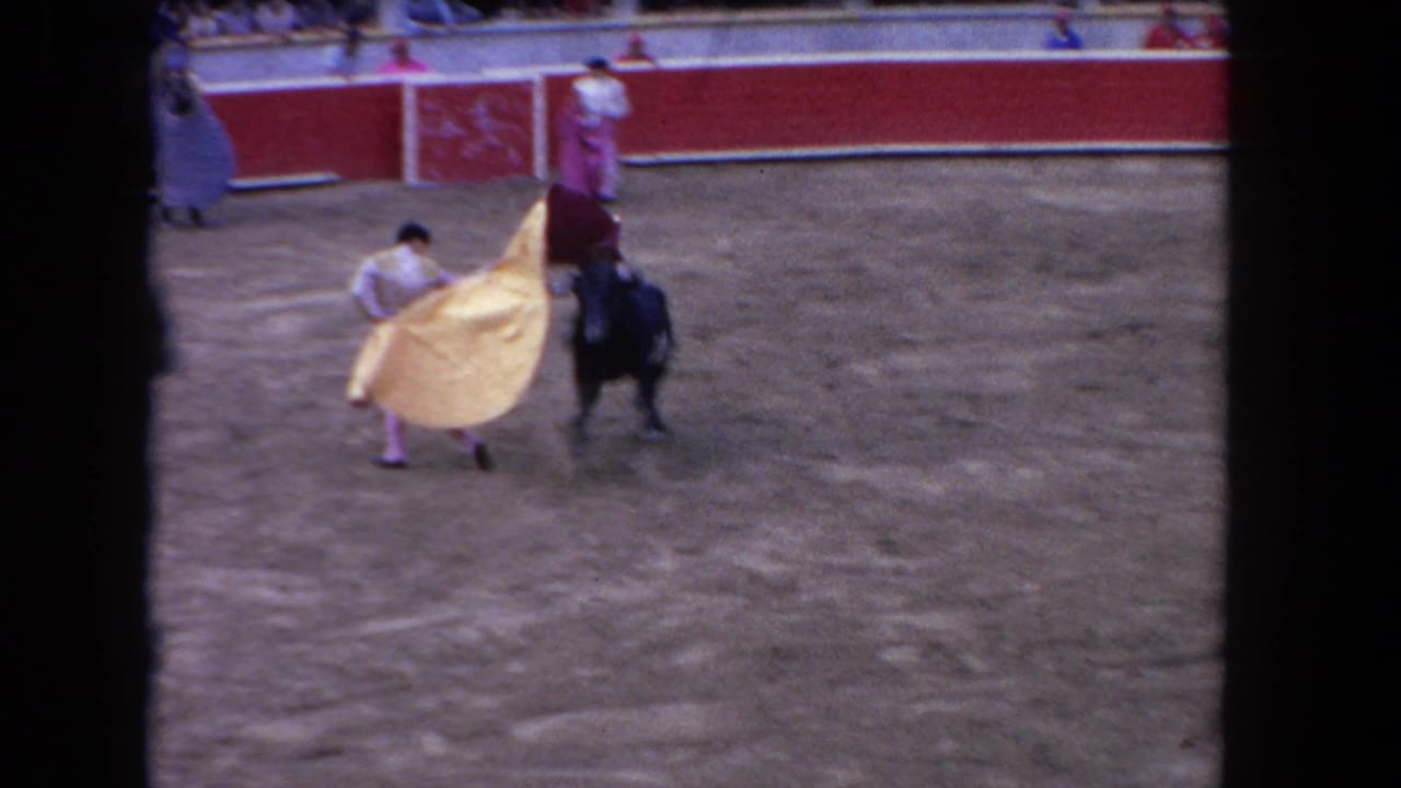 Matador sweeps a large golden-yellow cape toward a dark bull; red arena barrier wall visible in background.