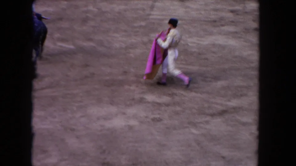 8mm home-movie frame, c.1962 Los Angeles: matador in white suit walks with pink cape across arena sand; red barrera wall with bourbon whiskey billboard and spectators behind.