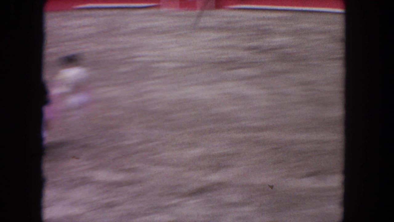 8mm home-movie frame, c.1962 Los Angeles: motion-blurred matador with pink cape in mid-arena action; bourbon whiskey billboard and red barrera wall visible in background.
