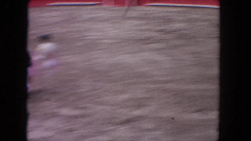 8mm home-movie frame, c.1962 Los Angeles: motion-blurred matador with pink cape in mid-arena action; bourbon whiskey billboard and red barrera wall visible in background.