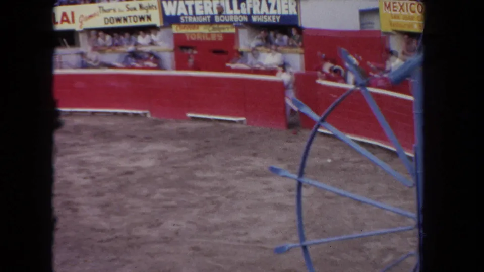 8mm home-movie frame, c.1962 Los Angeles: end-of-clip arena composition showing red barrera, bourbon whiskey sign, partial Mexico venue sign, spectators, and blue railing obscuring foreground.