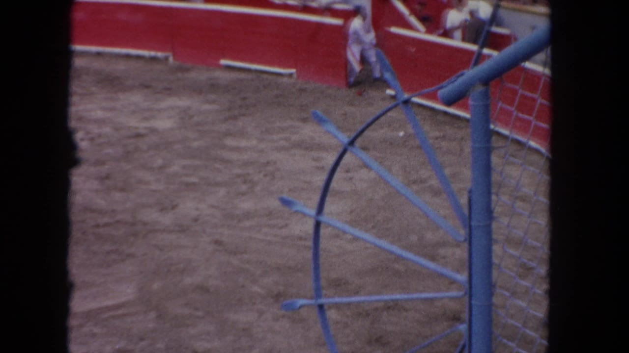 8mm color frame circa 1962: wide bullring interior with open sandy floor, curved red barrera walls, blue metal gate structure at right, single figure near far barrier.