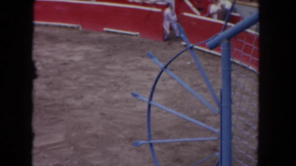 8mm color frame circa 1962: wide bullring interior with open sandy floor, curved red barrera walls, blue metal gate structure at right, single figure near far barrier.