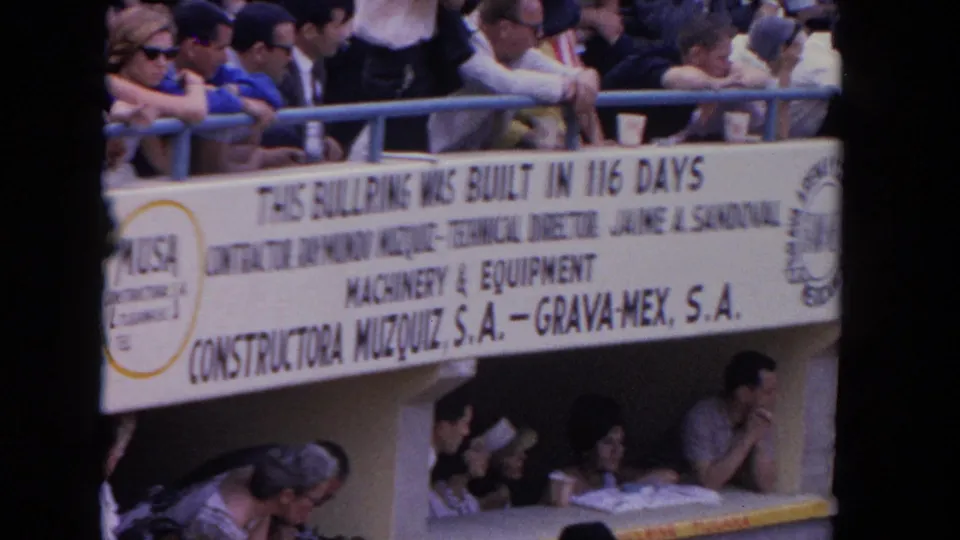 8mm color frame from 1962 showing crowd seating at a Los Angeles temporary bullfighting arena, with the Constructora Muzquiz S.A. credit banner prominent and a lower seated section visible.