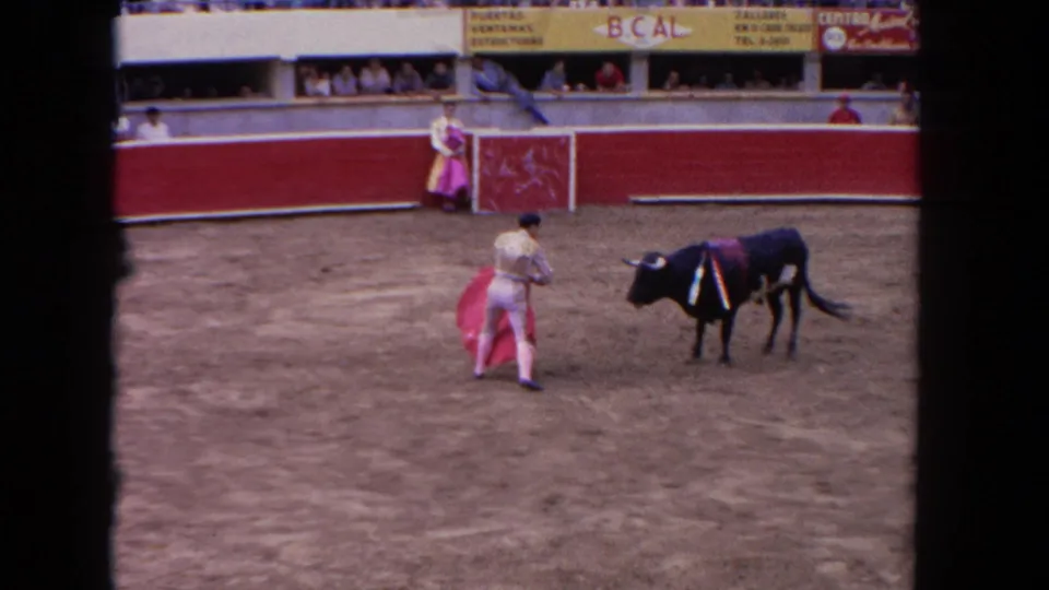 Bullfighting arena circa 1962; matador uses a pink muleta cape against a dark bull; second torero at left; arena barriers display Tijuana-area advertising.