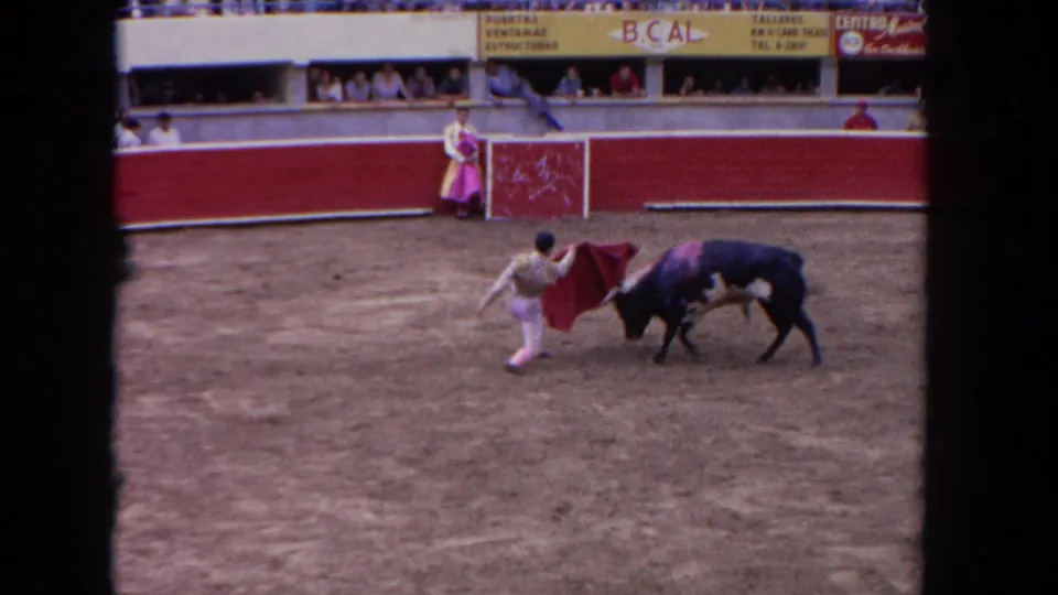 8mm home movie frame circa 1962 showing a matador in white-and-pink traje de luces working a red cape against a dark bull in a mid-century bullfighting arena.