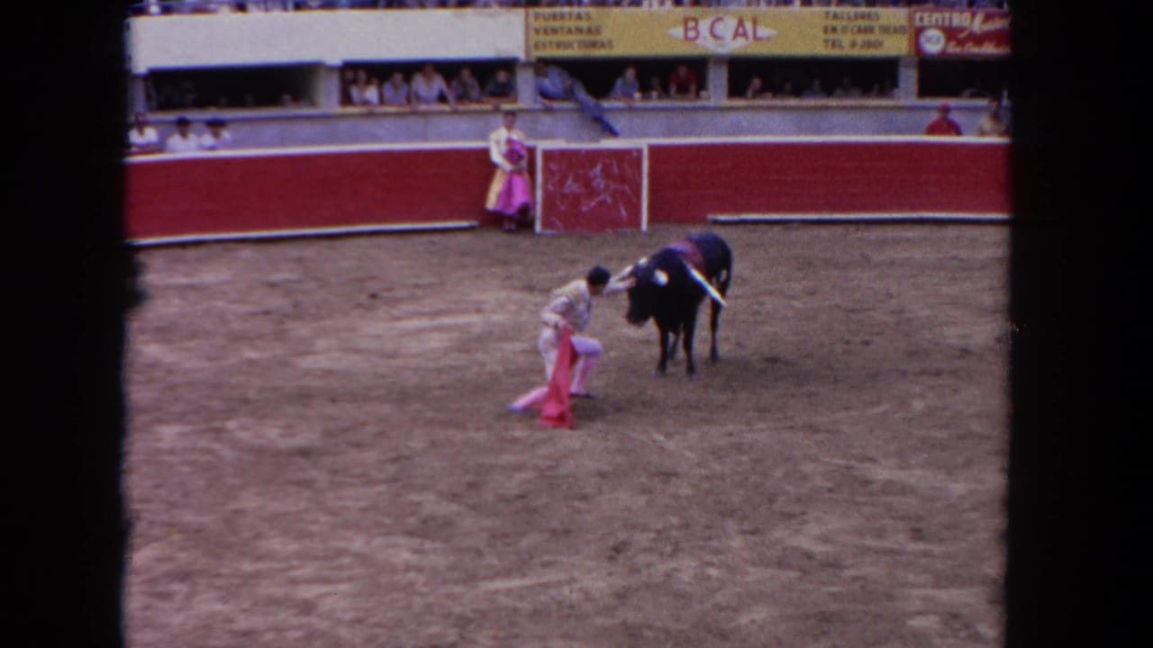 8mm color frame circa 1962 showing a bull charging toward a matador, with arena advertising banners including 'B.CAL' text visible in the background stands.
