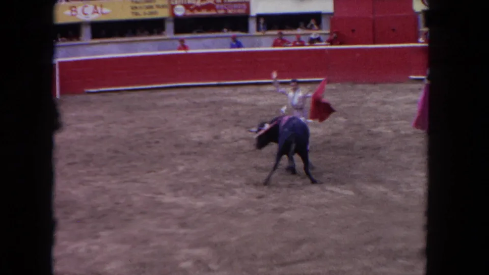 8mm home movie circa 1962 of a matador performing in mid-arena with a red cape while a dark bull charges; partial pink-costumed figure at frame edge, B.CAL banners visible in stands.