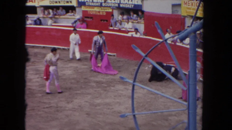 8mm frame c. 1962 showing matadors in magenta uniforms in a bullring, with a decorative blue circular metal gate at right and advertising signage including a 'MEXICO' banner above the spectator stands.