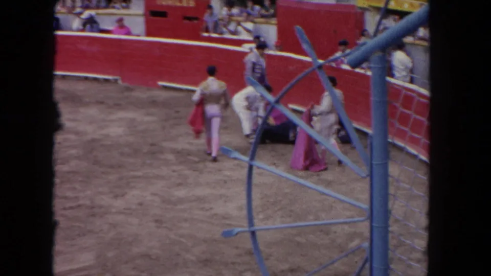 1962 8mm home-movie view from spectator stands of a Los Angeles bullfight arena; matadors in trajes de luces move near the curved red barrera.