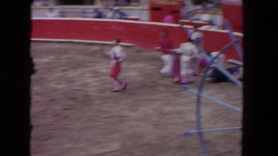 Motion-blurred 8mm frame of matadors running in a 1962 Los Angeles bullfight arena, shot handheld from spectator stands through foreground poles.