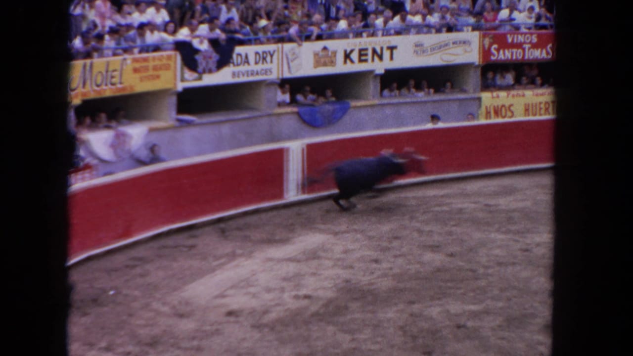 Wide 8mm frame from a 1962 bullfight showing a dark bull near the red circular barrera; Kent cigarette, Vinos Santo Tomas, and Canada Dry Beverages banners line the stands.