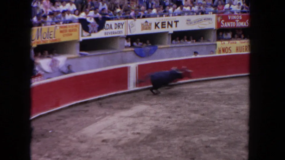 Wide 8mm frame from a 1962 bullfight showing a dark bull near the red circular barrera; Kent cigarette, Vinos Santo Tomas, and Canada Dry Beverages banners line the stands.