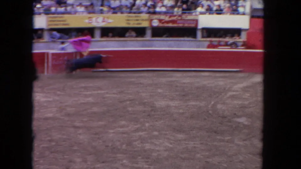 8mm home-movie frame circa 1962 showing a matador with a pink cape facing a dark bull near the red barrera; packed spectator stands and advertising hoardings visible above.