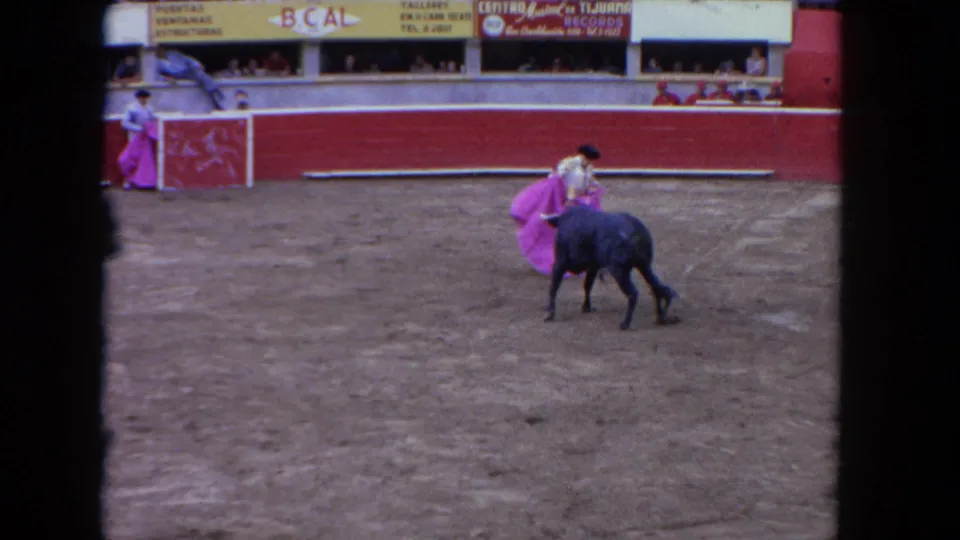 8mm color frame, c.1962: matador in white suit holds pink cape alongside dark bull in sunlit arena; advertising boards visible in upper stands.