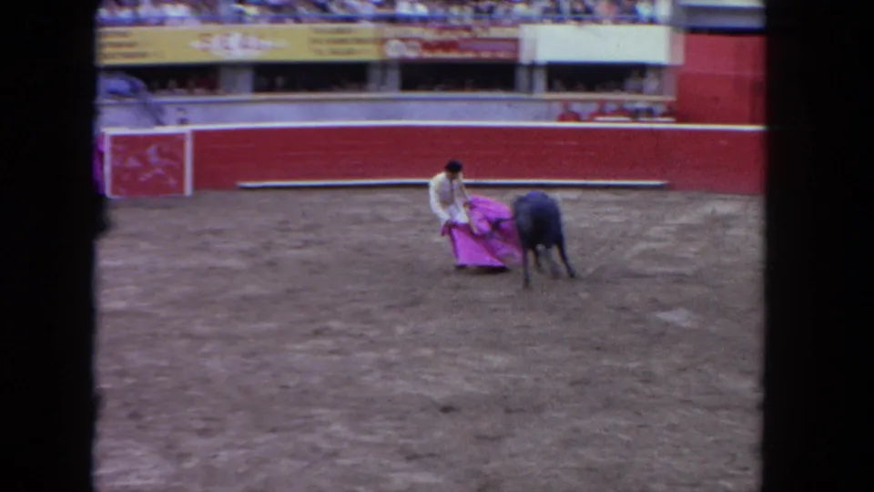 8mm home-movie frame, c.1962: matador in low crouch extends muleta to draw bull past; red barrera and tiered spectator stands visible behind.