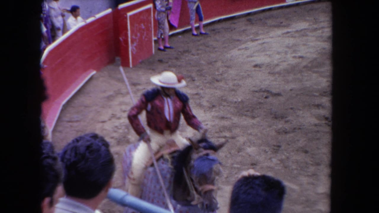 Picador on horseback near red barrier with a bull at lower right, colorful advertising boards on callejón wall, spectator heads in foreground.