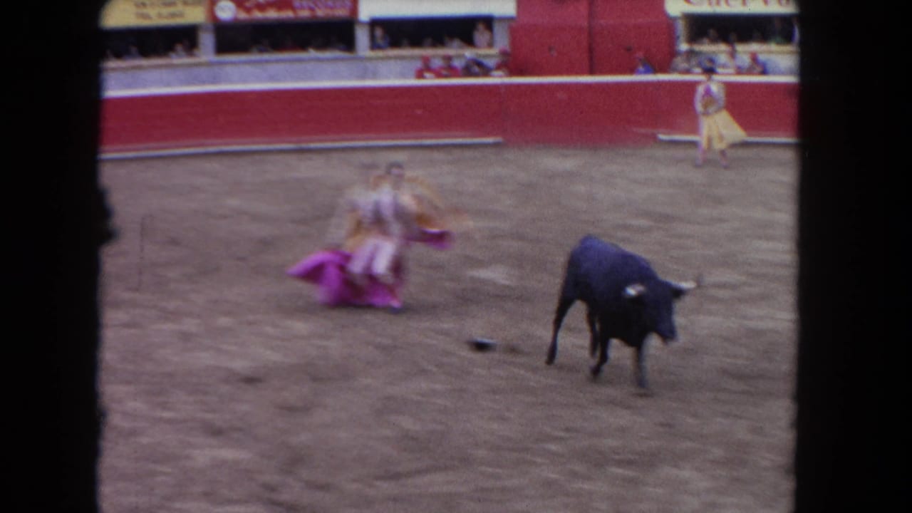 8mm footage from 1962, likely Los Angeles: matador in pink costume faces a black bull in a dirt arena with red barrier walls, spectators, and advertising banners in the background.