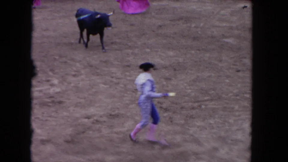 1960s color 8mm frame of a matador in light-blue traje de luces crossing a bullring, black bull at distance, pink cape at frame edge