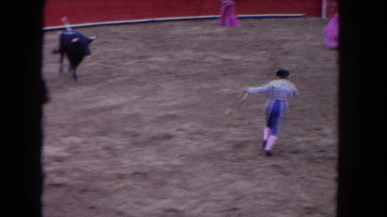 1960s color 8mm frame showing matador's back and black bull at distance in a sun-lit bullring, pink cape visible at edge