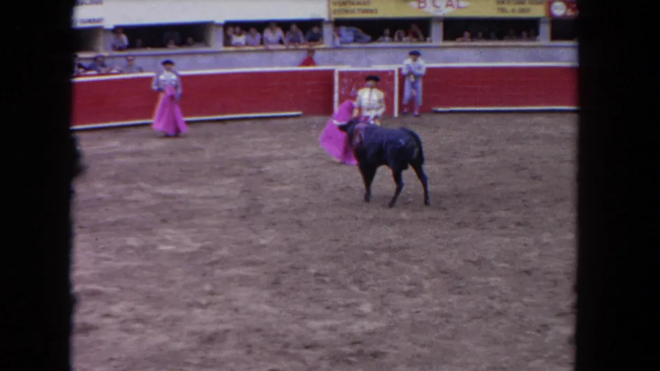 1960s 8mm color frame: matador with pink cape and black bull near red barrera, packed spectator stands and advertising signage visible in background