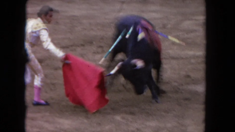 1960s 8mm close-up of a bullfight cape pass — matador in traje de luces holds a red muleta as a dark bull with colorful banderillas charges across a sandy arena.