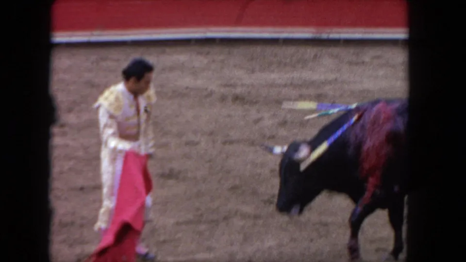 1960s 8mm medium shot of a matador in traje de luces holding a red muleta low as a banderilla-adorned dark bull stands at close range in a sandy arena.