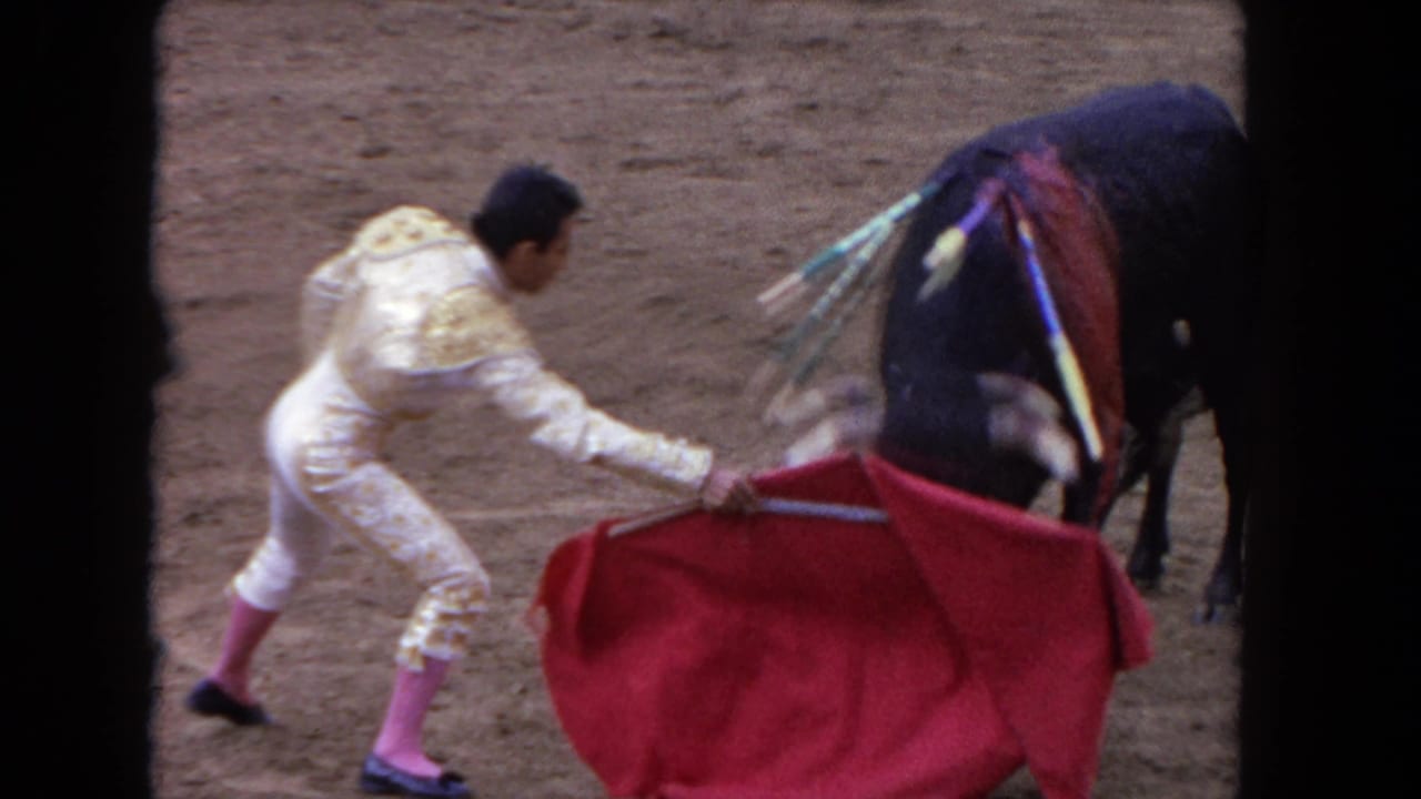 8mm close-up from circa 1962: matador in embroidered traje de luces sweeping the muleta as a black bull bearing colorful banderillas passes close.