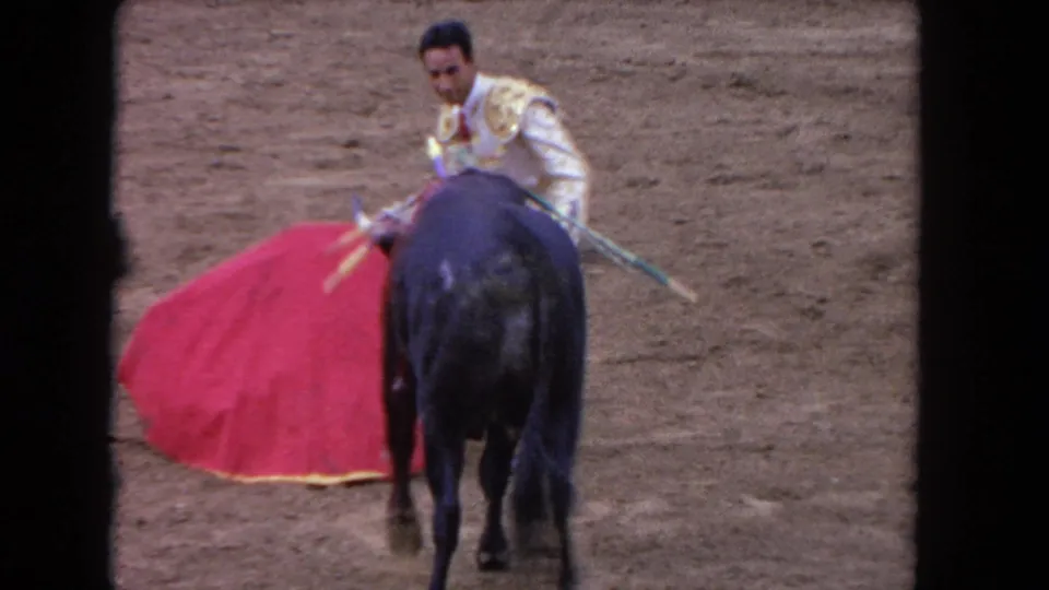 8mm shot from behind showing a matador in white-gold embroidered suit confronting a black bull bearing banderillas head-on in a sandy arena, circa 1962.