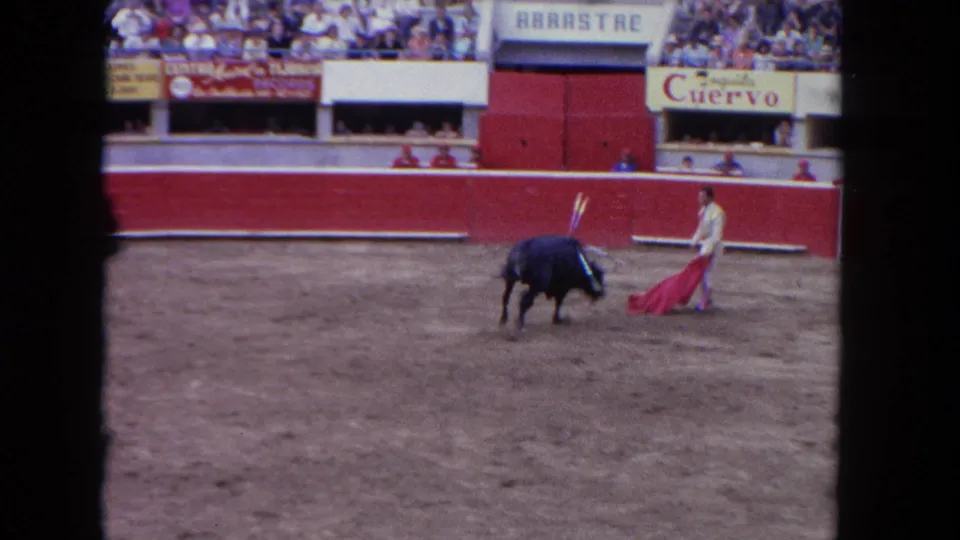 Wide-angle 8mm frame from circa 1962 showing a bullfighting arena with crowd in the stands, red barrera wall, matador and bull at center, and legible Cuervo and ARRASTRE signage.