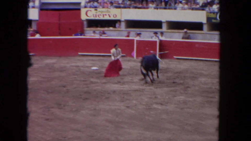 1960s corrida close view: matador in light traje de luces holding red muleta toward approaching bull, ring attendants at barrera, Tequila Cuervo banner prominent.