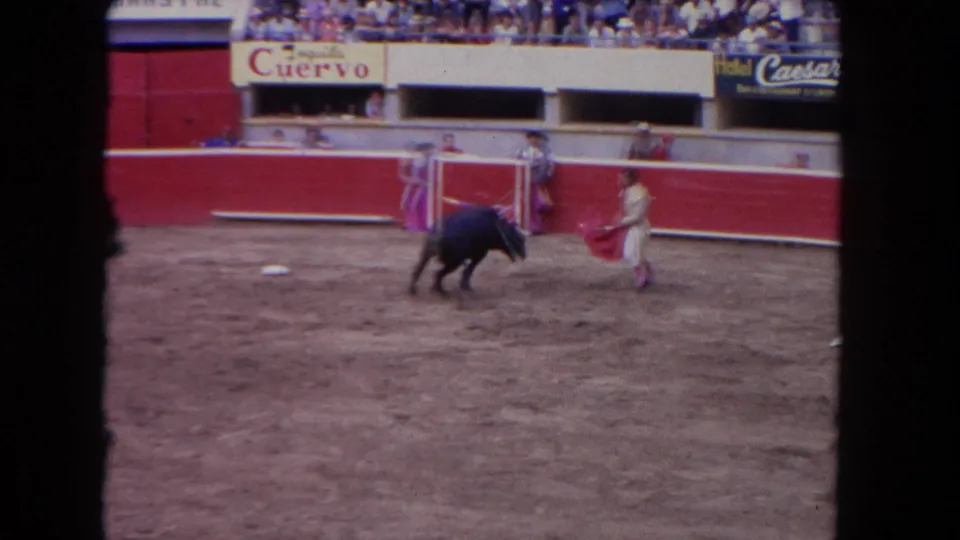 1960s corrida: multiple toreros and bull near red barrera, Tequila Cuervo banner and second partially legible sponsor sign above packed bleachers.