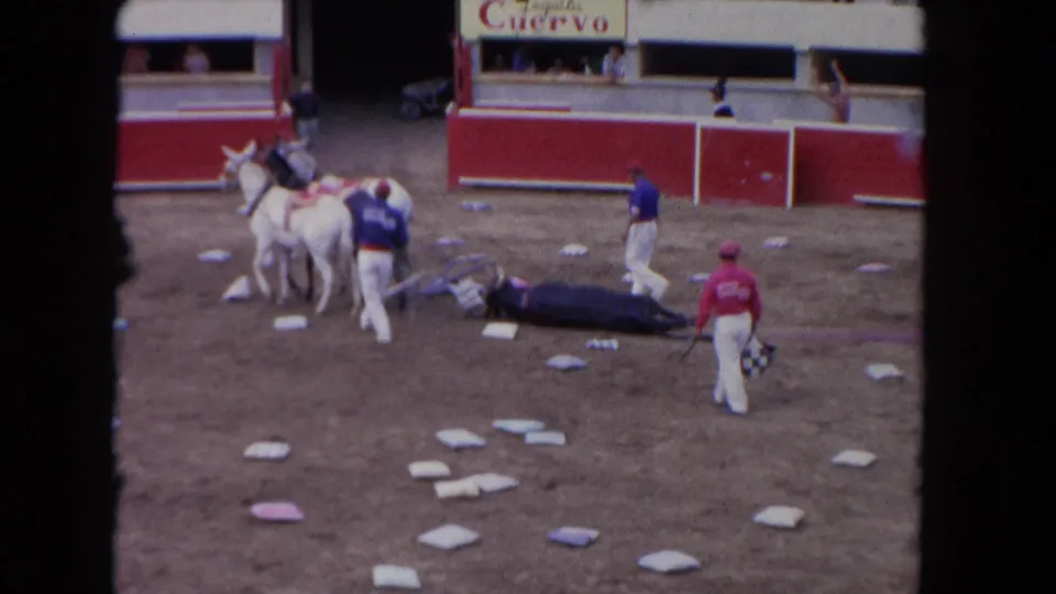 1962 Los Angeles 8mm frame showing attendants and mounted riders near an injured bull in a dirt arena with spectators behind red barriers.