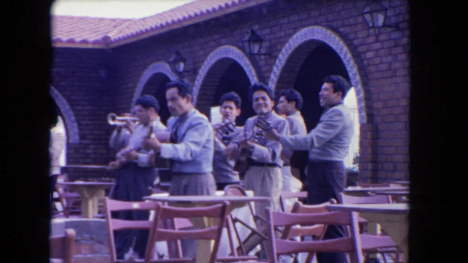 1962 Los Angeles home-movie frame showing musicians with trumpet, guitar, and clapping near brick arches and courtyard seating.