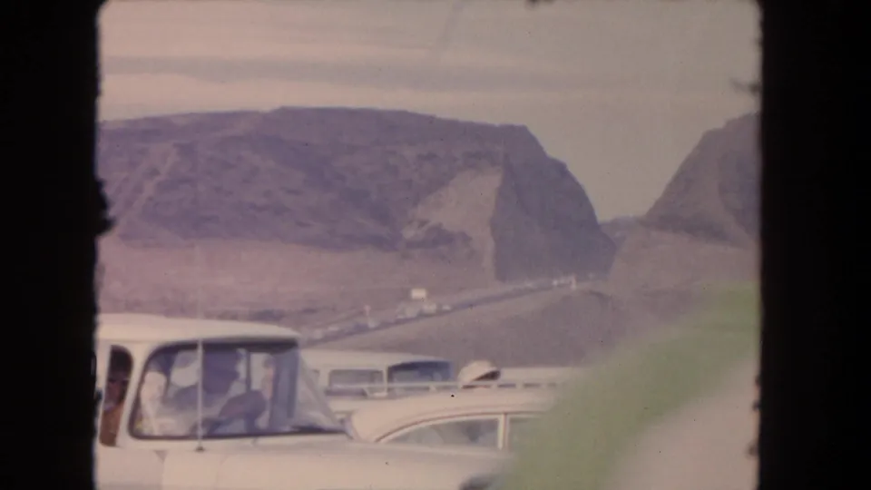 1962 8mm home-movie frame of classic cars moving through traffic with arid mountains in the background.
