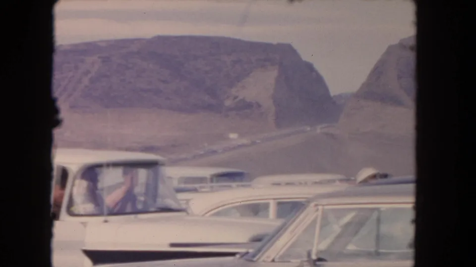 1962 California road scene with a white convertible and other classic cars against a desert mountain backdrop.