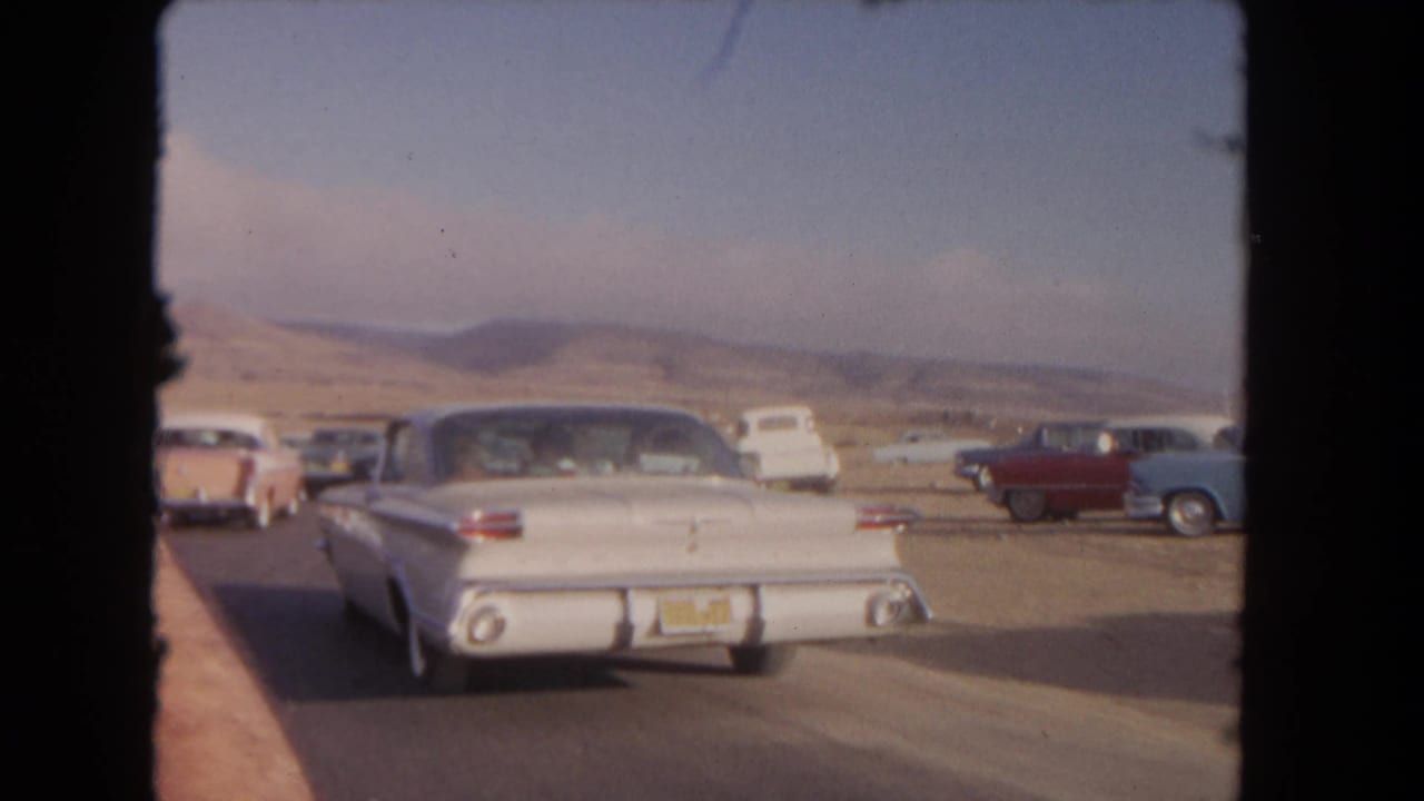 Rear view of a white sedan on a desert road with parked cars nearby.