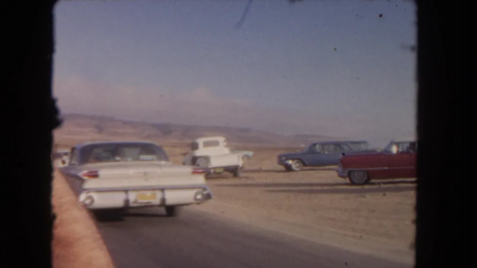 The camera follows a white sedan from behind on a desert road, with mid-century cars parked along the roadside and dry hills in the background.