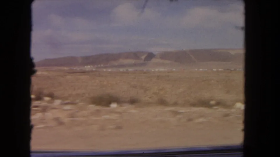 Moving 8mm view across open desert ground toward low hills near Los Angeles, with motion blur and a dark window or frame edge at the side.