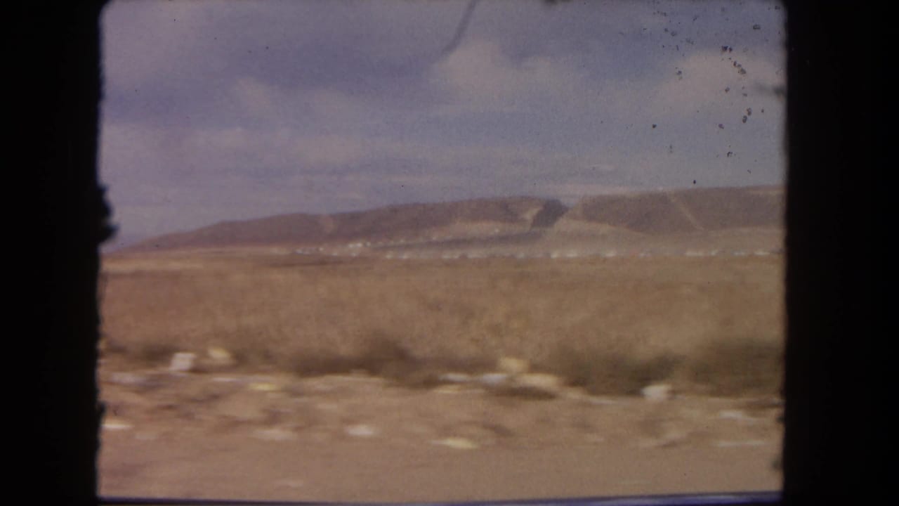 1962 8mm car-window view of arid terrain, scrub, pale sky, and distant low hills near Los Angeles, California.