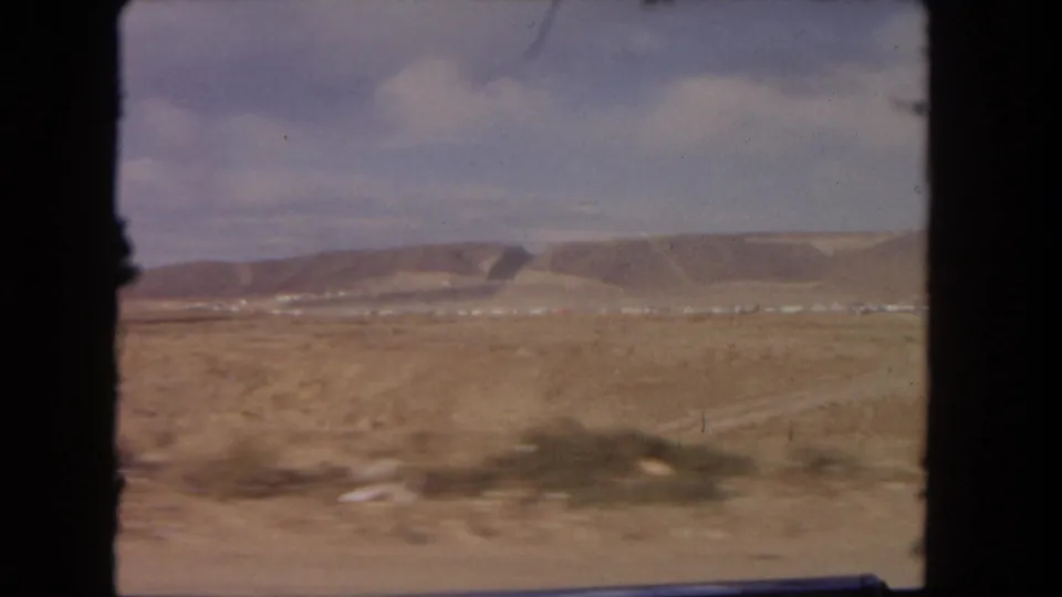 1962 8mm view from a moving vehicle across dry plains toward low hills, with small distant vehicles near the road.