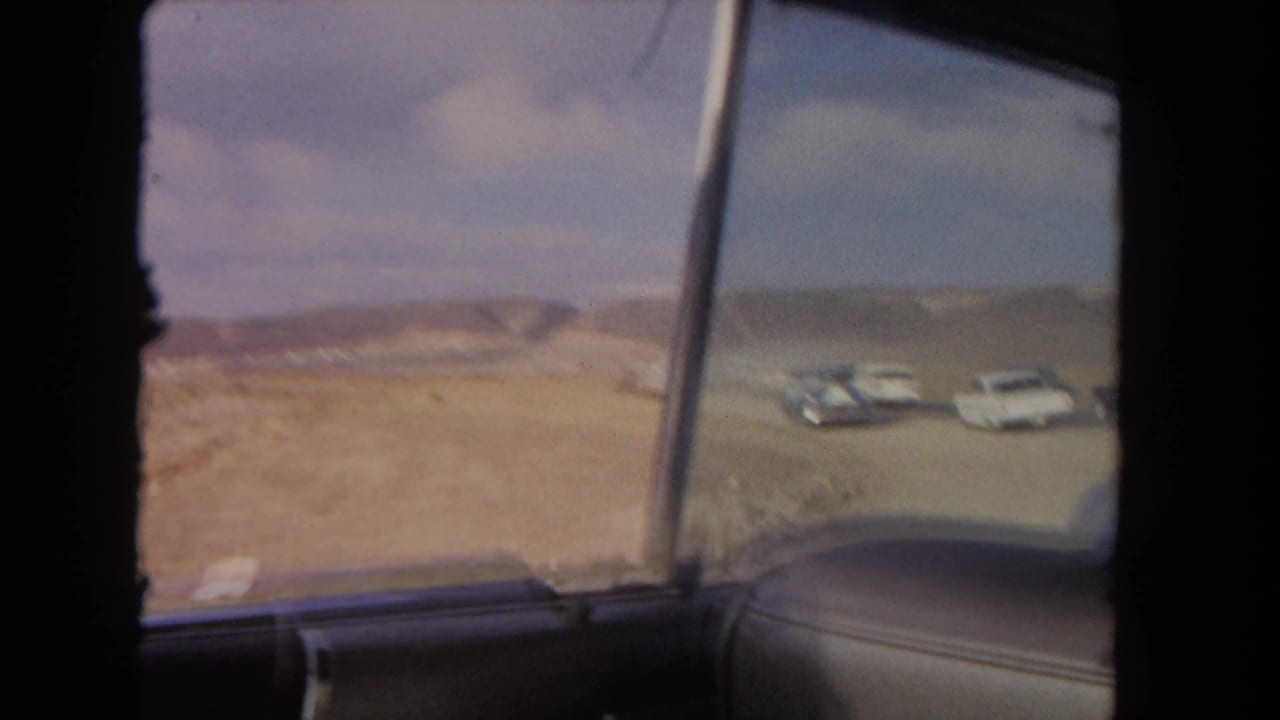 View through a car side window toward dusty hills and classic cars on open ground.