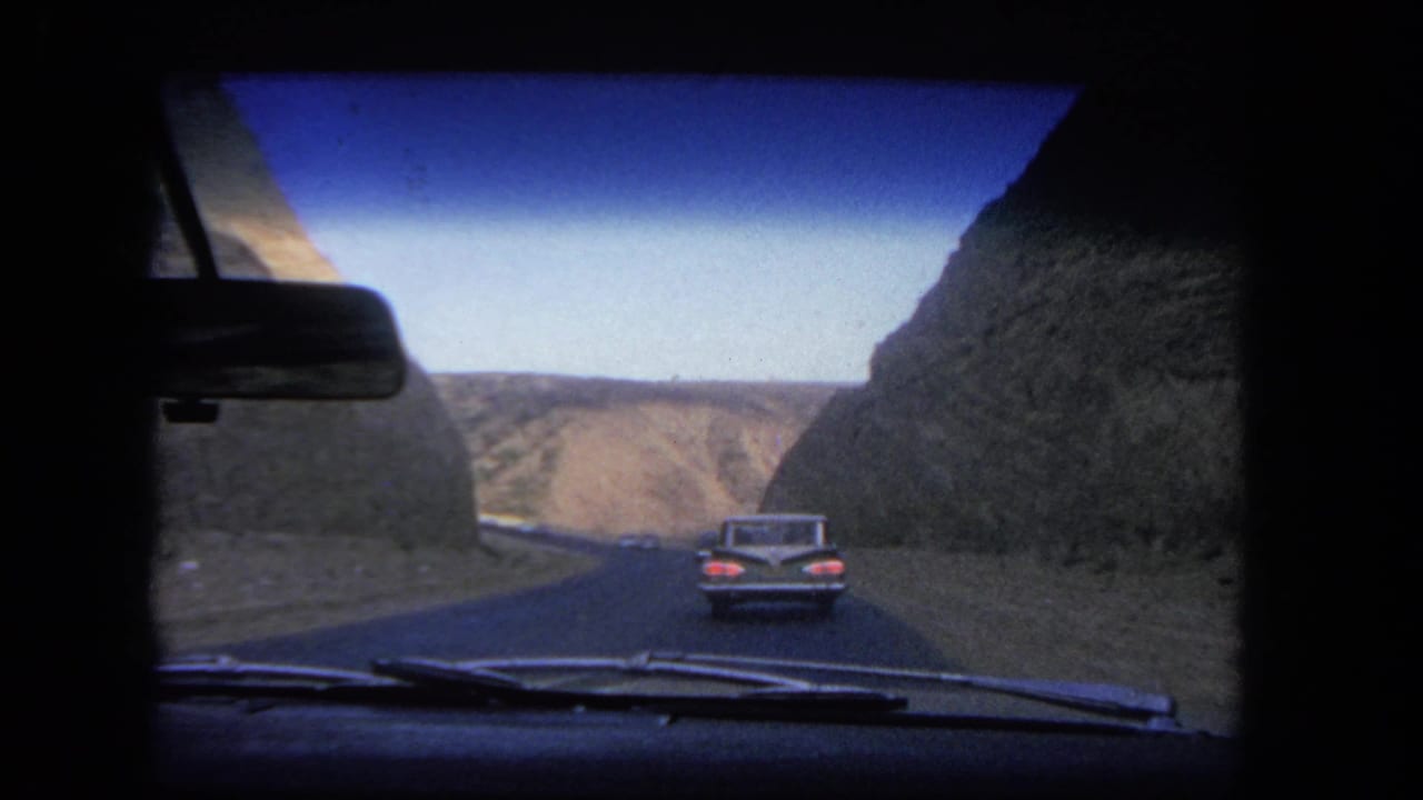 1962 Los Angeles-area driving view through a windshield, following a classic car along a road cut between arid hills.