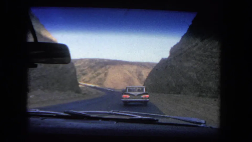 Home-movie passenger view from a moving car in 1962, with a classic car ahead on a road through dry Los Angeles hills.