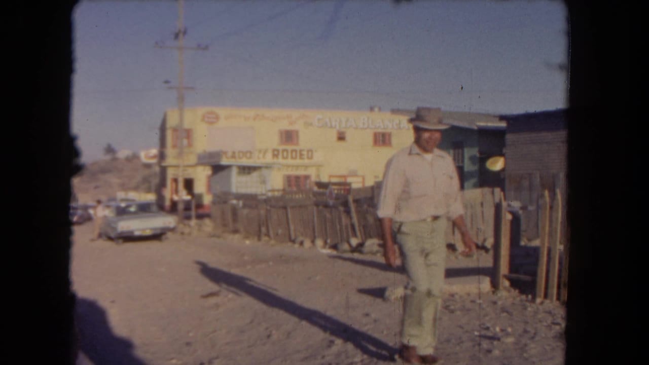 Man in a cowboy hat walking beside a dirt road near a yellow building with signage.