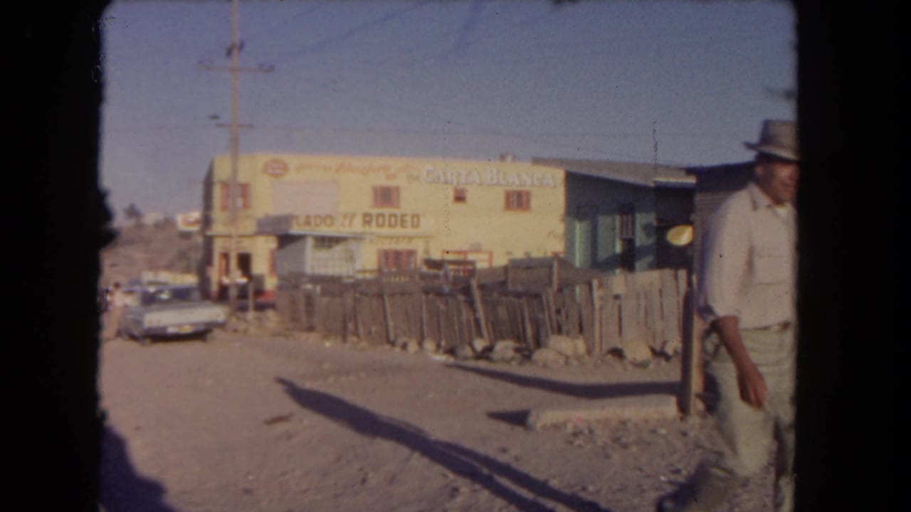 1962 Los Angeles 8mm frame showing the man partly at the right edge as he passes a rough fence, dirt road, parked car, and yellow building with commercial signage.